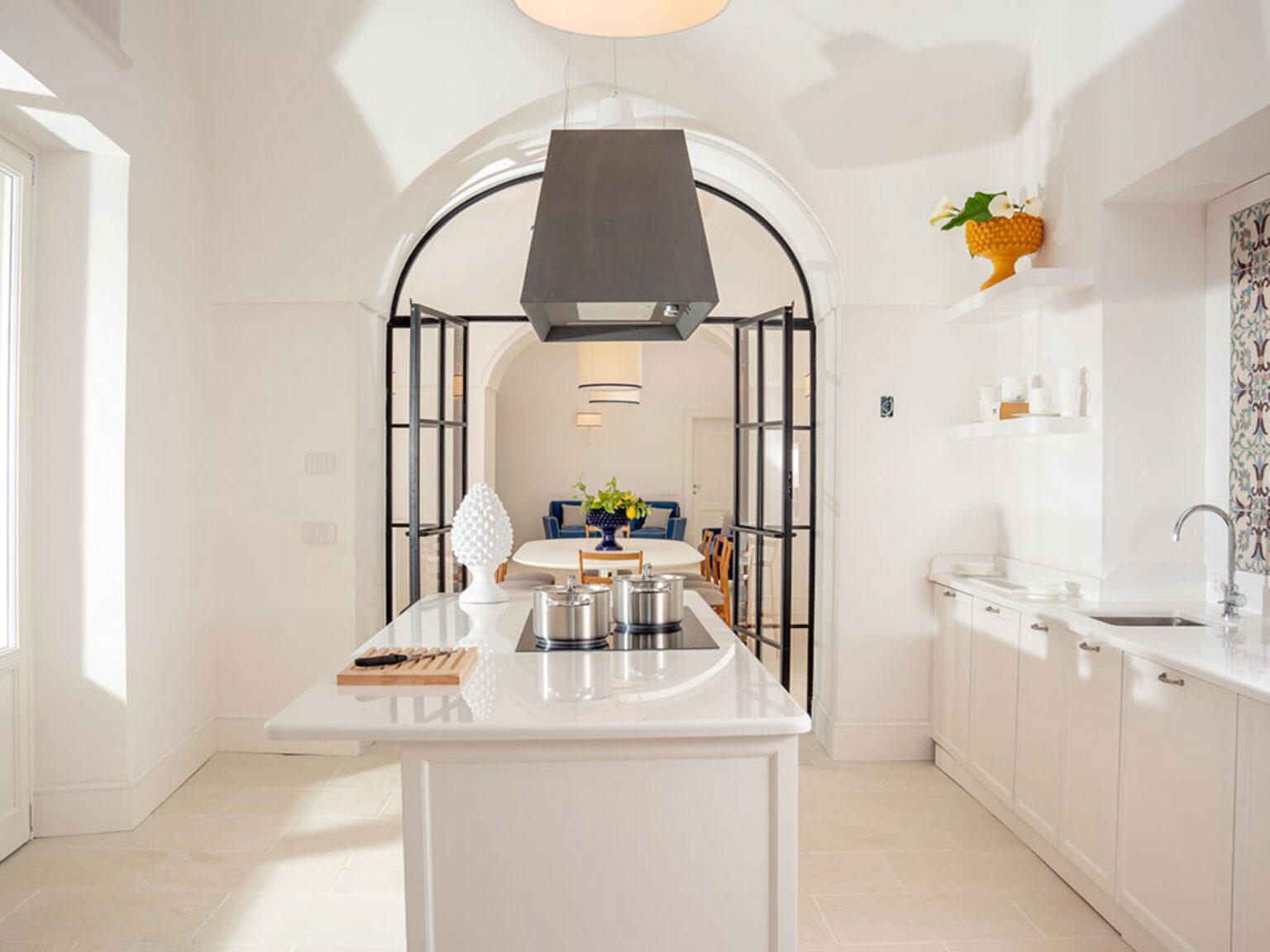 Modern kitchen featuring a spacious island, sleek white cabinetry, and a stylish black hood. The open layout includes decorative elements like fresh flowers and vibrant accents, creating a bright and inviting atmosphere. | Only on SopranoVillas