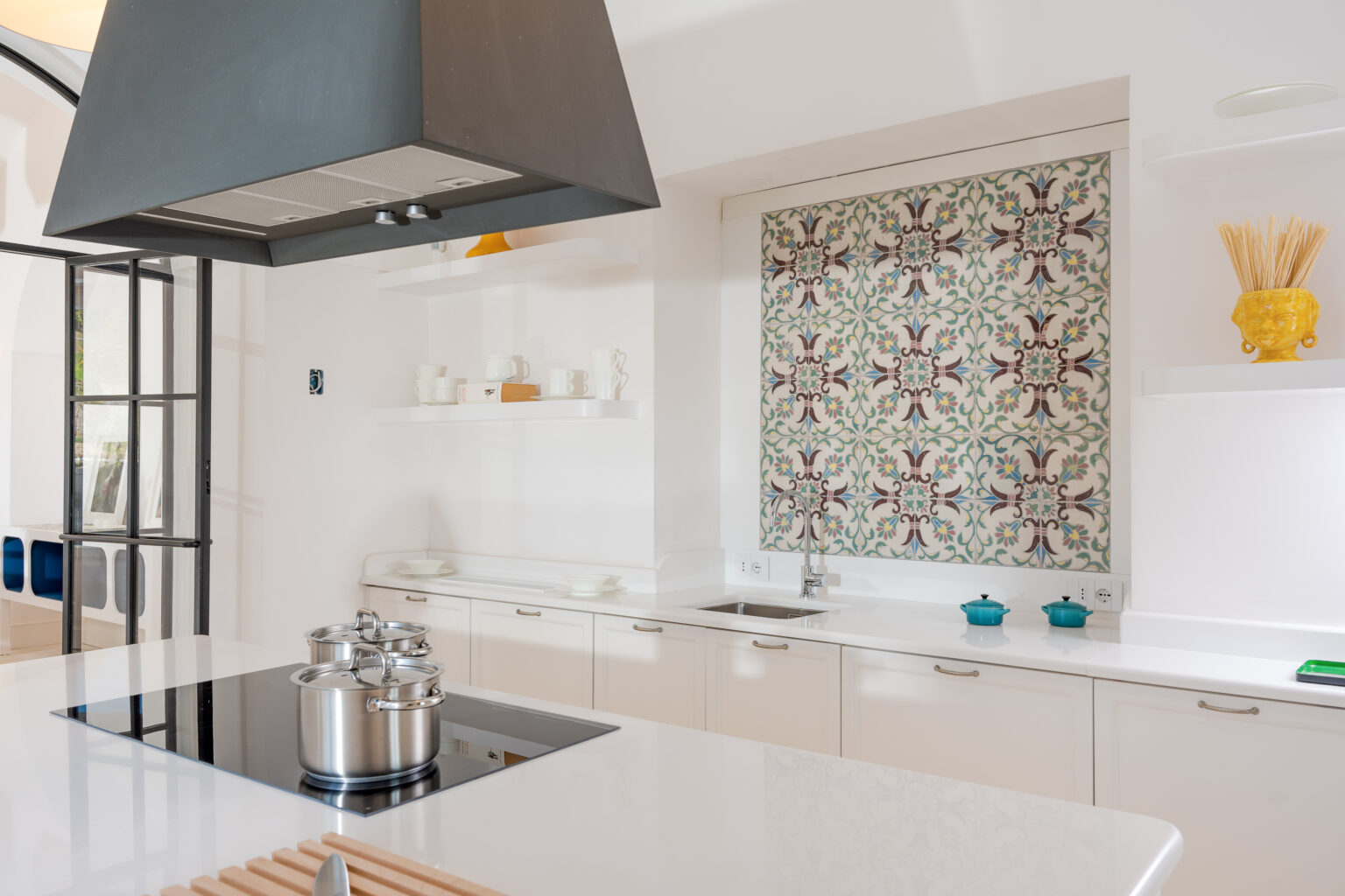 Modern kitchen interior featuring a sleek white countertop, stainless steel pots on an induction cooktop, and a decorative patterned backsplash. Open shelving displays minimalist dishware and a vibrant yellow vase, enhancing the contemporary design. | Only on SopranoVillas