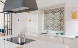 Modern kitchen interior featuring a sleek white countertop, stainless steel pots on an induction cooktop, and a decorative patterned backsplash. Open shelving displays minimalist dishware and a vibrant yellow vase, enhancing the contemporary design.
