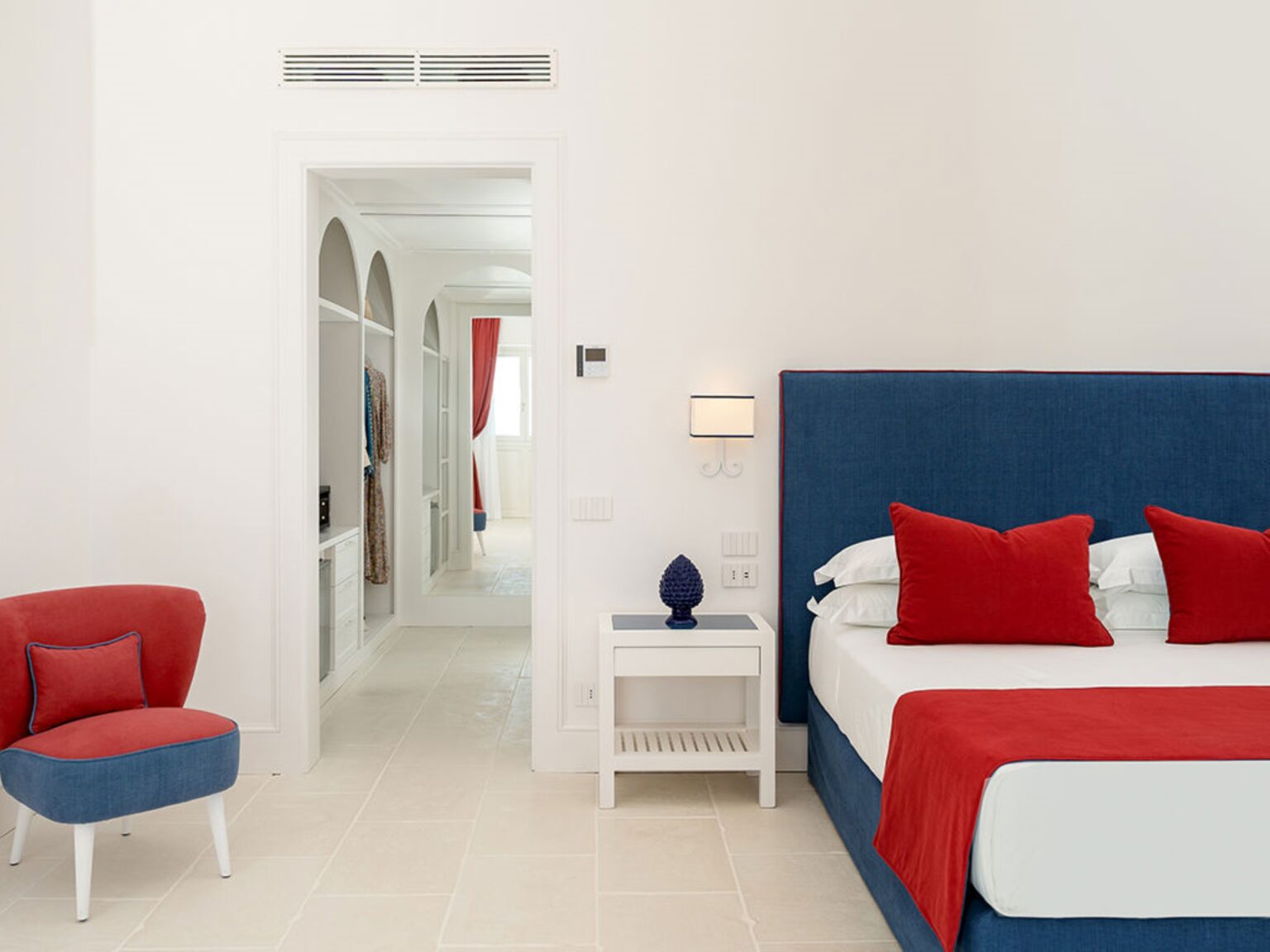 Modern hotel room featuring a blue and white color scheme, a comfortable bed with red accent pillows, a stylish red chair, and an open doorway leading to a bright, airy hallway. Ideal for travelers seeking contemporary accommodations. | Only on SopranoVillas