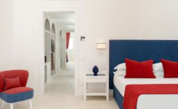 Modern hotel room featuring a blue and white color scheme, a comfortable bed with red accent pillows, a stylish red chair, and an open doorway leading to a bright, airy hallway. Ideal for travelers seeking contemporary accommodations.