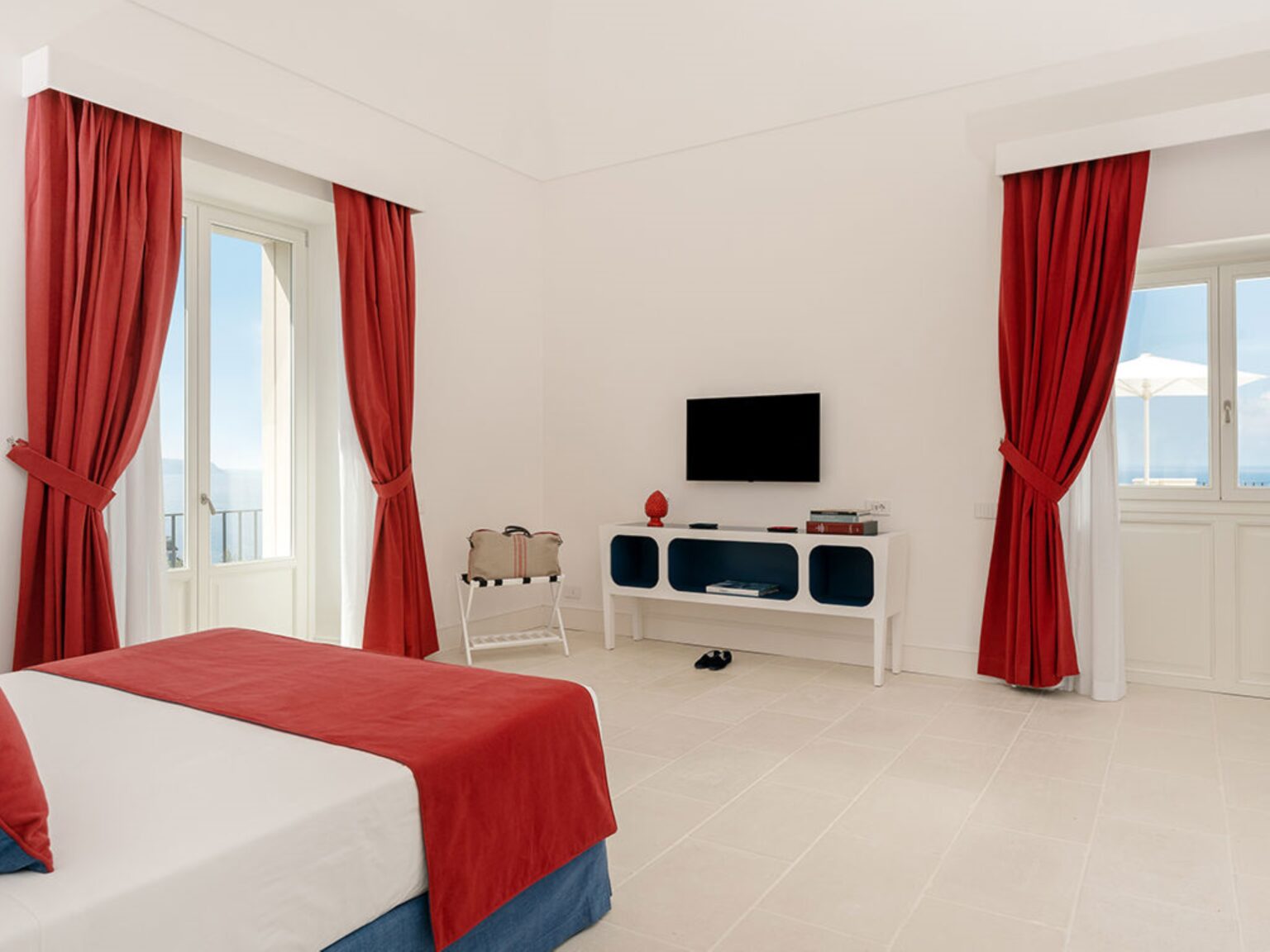 Bright and airy hotel room featuring a red and white color scheme, large windows with sheer curtains, a flat-screen TV, and modern furnishings, ideal for a comfortable stay. | Only on SopranoVillas
