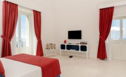 Bright and airy hotel room featuring a red and white color scheme, large windows with sheer curtains, a flat-screen TV, and modern furnishings, ideal for a comfortable stay.