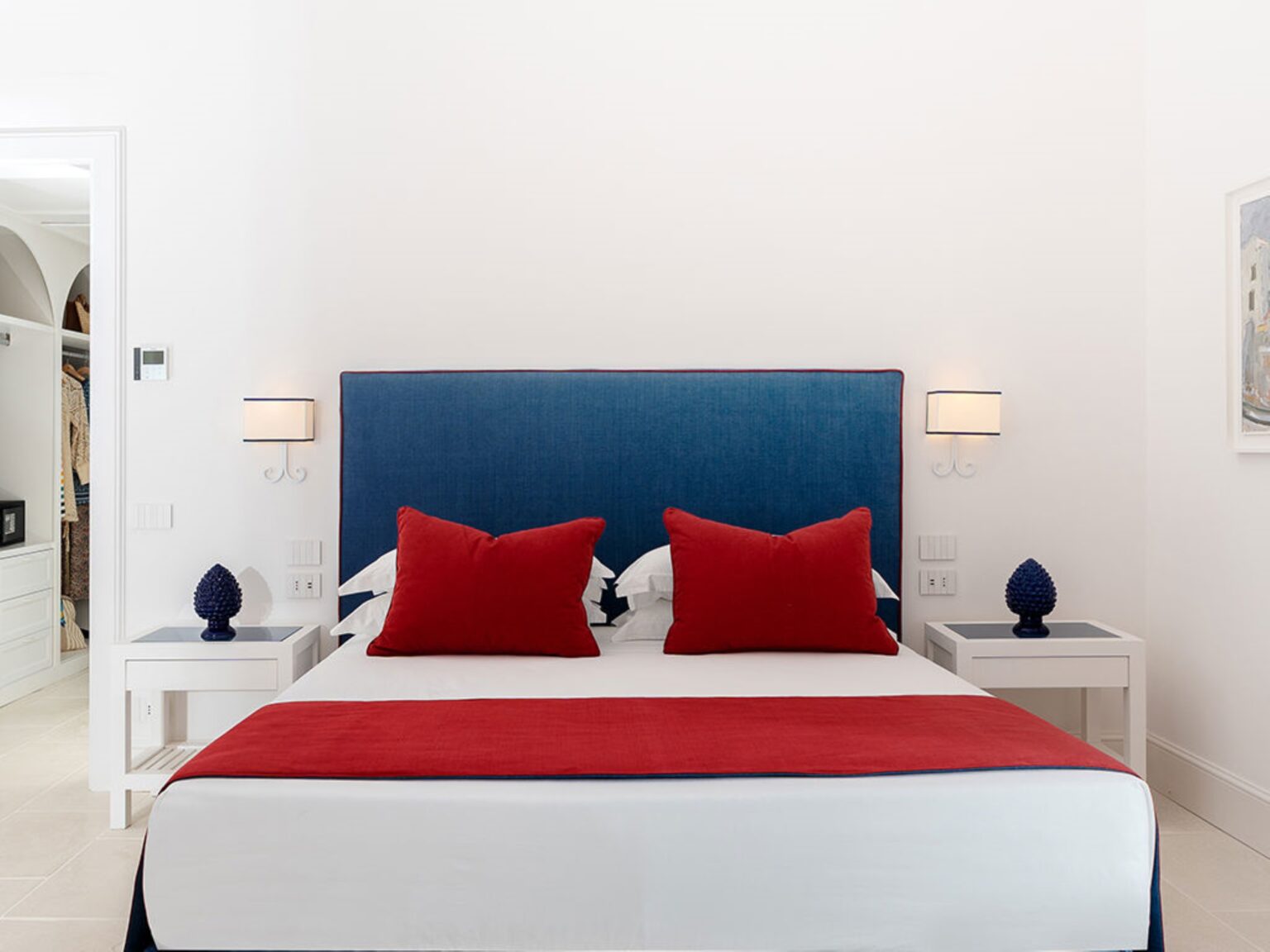 Modern bedroom featuring a blue upholstered headboard, red decorative pillows, and white nightstands with blue lamps, creating a stylish and inviting atmosphere. | Only on SopranoVillas