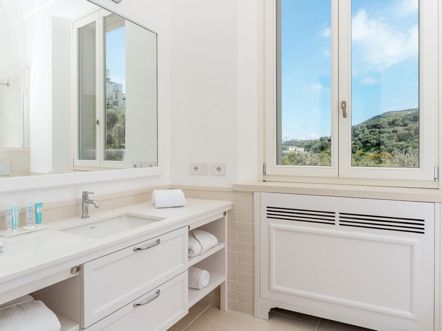 Bright and modern bathroom featuring a white vanity with a mirror, neatly arranged towels, and a large window offering a view of green hills and a blue sky. | Only on SopranoVillas