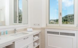 Bright and modern bathroom featuring a white vanity with a mirror, neatly arranged towels, and a large window offering a view of green hills and a blue sky.