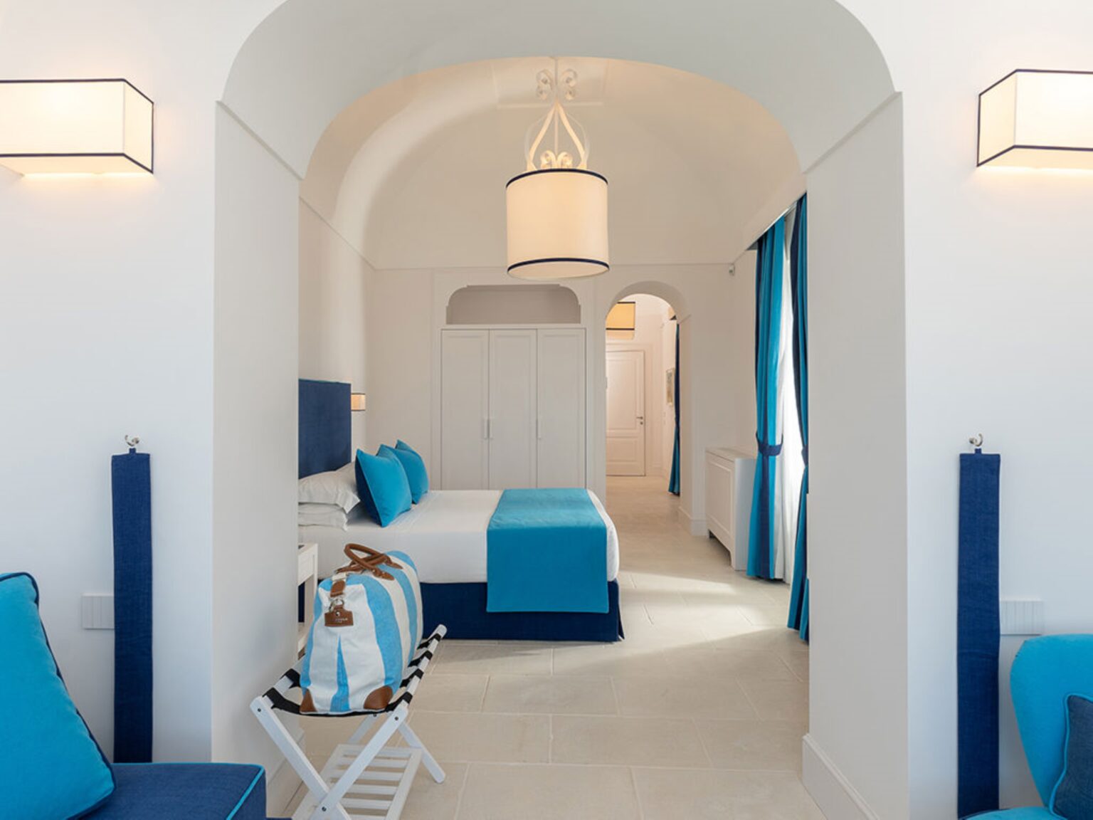 Modern hotel room with white walls and blue accents, featuring a queen-size bed, stylish light fixtures, and large windows with blue curtains. The room has a contemporary design, creating a bright and inviting atmosphere. Ideal for travelers seeking comfort and elegance. | Only on SopranoVillas