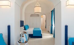 Modern hotel room with white walls and blue accents, featuring a queen-size bed, stylish light fixtures, and large windows with blue curtains. The room has a contemporary design, creating a bright and inviting atmosphere. Ideal for travelers seeking comfort and elegance.
