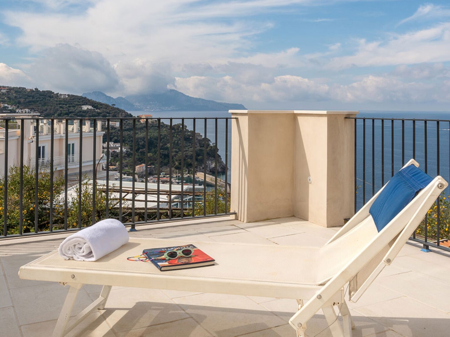 Lounge chair with a towel and sunglasses on a terrace overlooking a scenic coastal view, featuring mountains and a clear sky. Ideal for relaxation and enjoying the outdoors. | Only on SopranoVillas