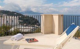 Lounge chair with a towel and sunglasses on a terrace overlooking a scenic coastal view, featuring mountains and a clear sky. Ideal for relaxation and enjoying the outdoors.
