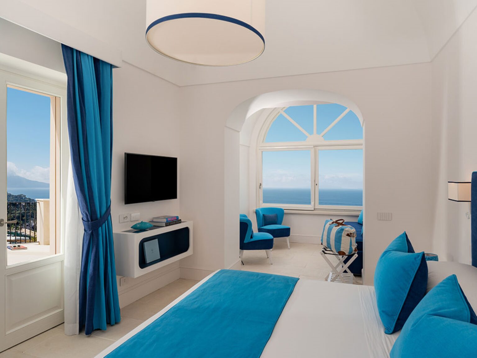 Bright and modern hotel room featuring blue accents, a large window with ocean views, a flat-screen TV, and comfortable seating. Ideal for a relaxing getaway. | Only on SopranoVillas