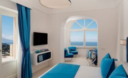 Bright and modern hotel room featuring blue accents, a large window with ocean views, a flat-screen TV, and comfortable seating. Ideal for a relaxing getaway.