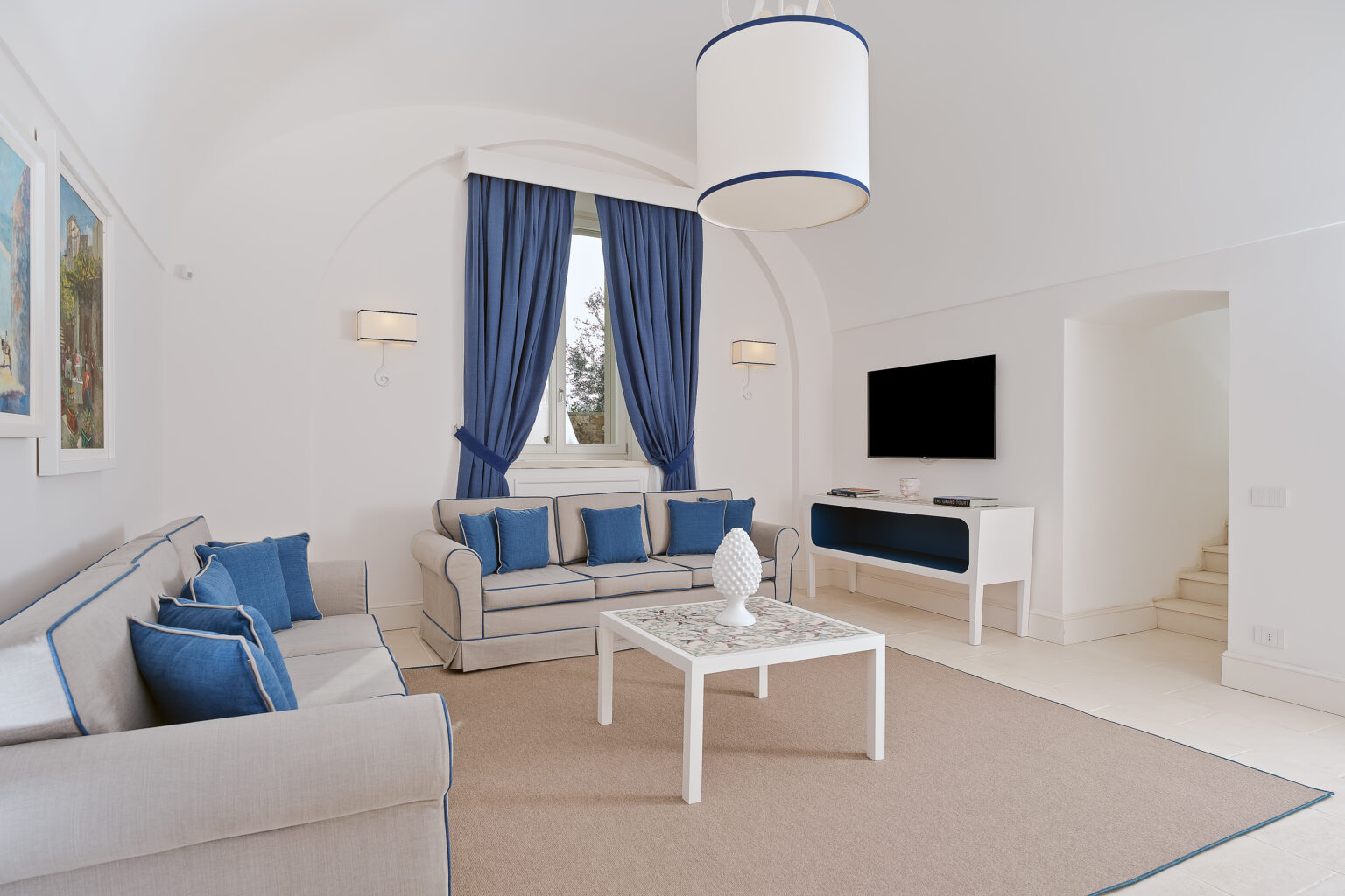 Bright and elegant living room featuring white walls, blue accents, and modern furnishings. The space includes a comfortable sofa, decorative cushions, a stylish coffee table, and large windows with blue curtains, creating a serene atmosphere. A flat-screen TV is mounted on the wall, enhancing the contemporary design. | Only on SopranoVillas