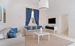 Bright and elegant living room featuring white walls, blue accents, and modern furnishings. The space includes a comfortable sofa, decorative cushions, a stylish coffee table, and large windows with blue curtains, creating a serene atmosphere. A flat-screen TV is mounted on the wall, enhancing the contemporary design.