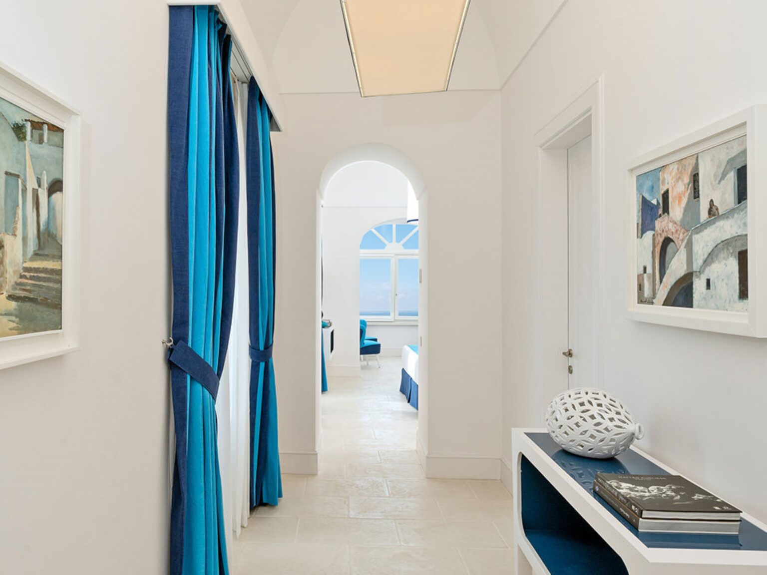 Bright and airy hallway featuring white walls, blue curtains, and framed artworks, leading to a sunlit room with arched windows. Modern decor accents the space, creating a serene atmosphere. | Only on SopranoVillas