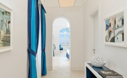 Bright and airy hallway featuring white walls, blue curtains, and framed artworks, leading to a sunlit room with arched windows. Modern decor accents the space, creating a serene atmosphere.