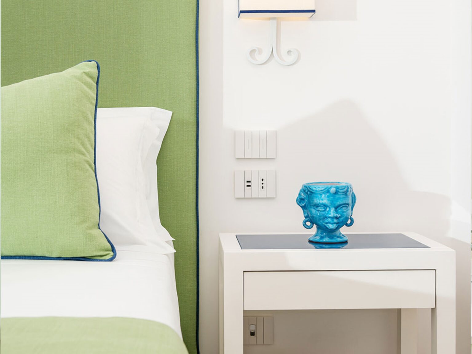 Bright and modern bedroom decor featuring a green accent wall, white bedding with a green and blue pillow, and a stylish blue decorative vase on a sleek white nightstand. The image captures a contemporary interior design aesthetic. | Only on SopranoVillas