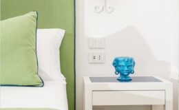 Bright and modern bedroom decor featuring a green accent wall, white bedding with a green and blue pillow, and a stylish blue decorative vase on a sleek white nightstand. The image captures a contemporary interior design aesthetic.