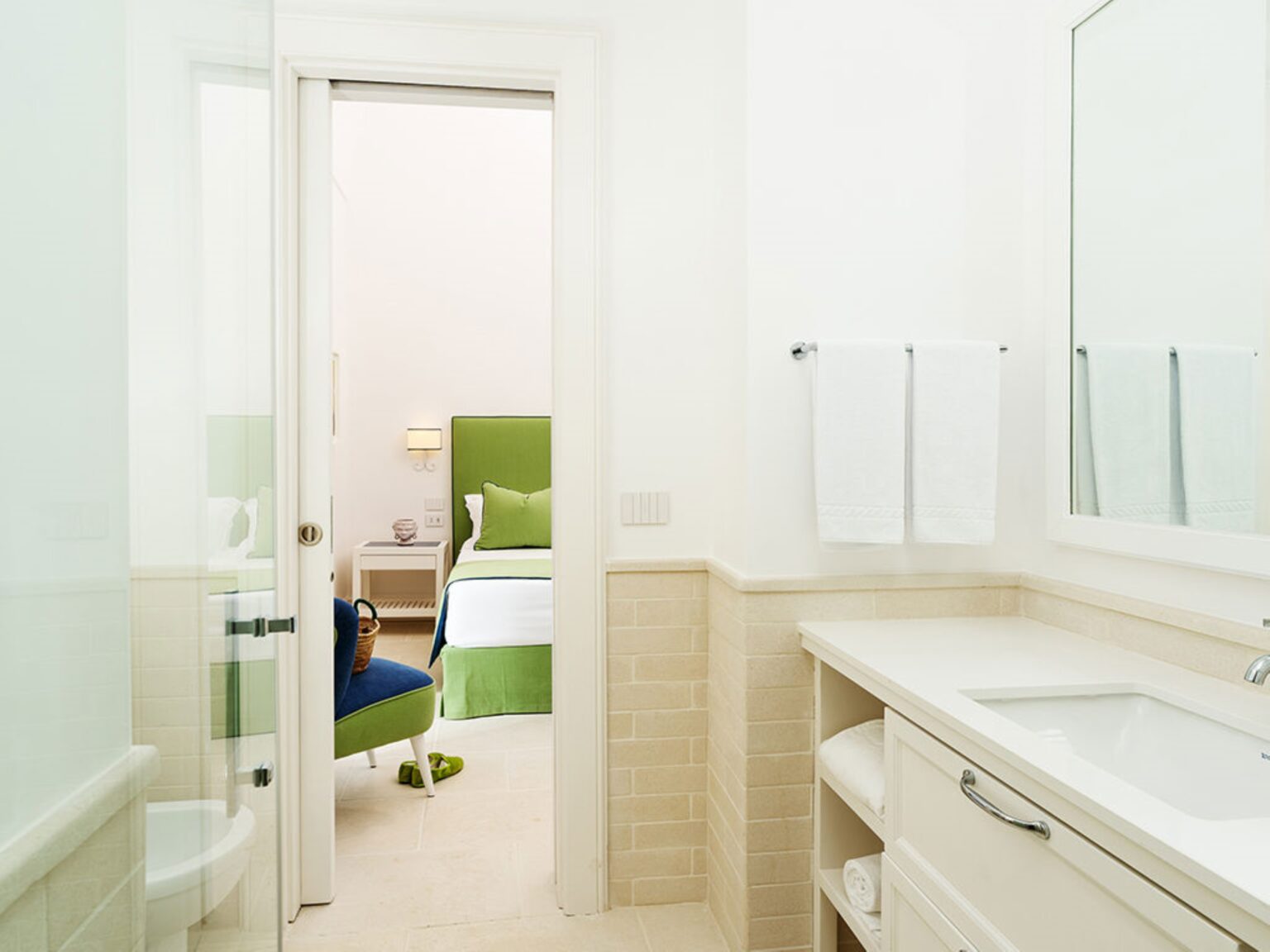 Modern bathroom interior with white walls and a sleek vanity, featuring a large mirror, towels hanging on the wall, and a view into a bright bedroom with green accents. | Only on SopranoVillas