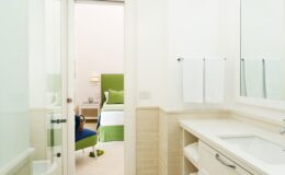 Modern bathroom interior with white walls and a sleek vanity, featuring a large mirror, towels hanging on the wall, and a view into a bright bedroom with green accents.