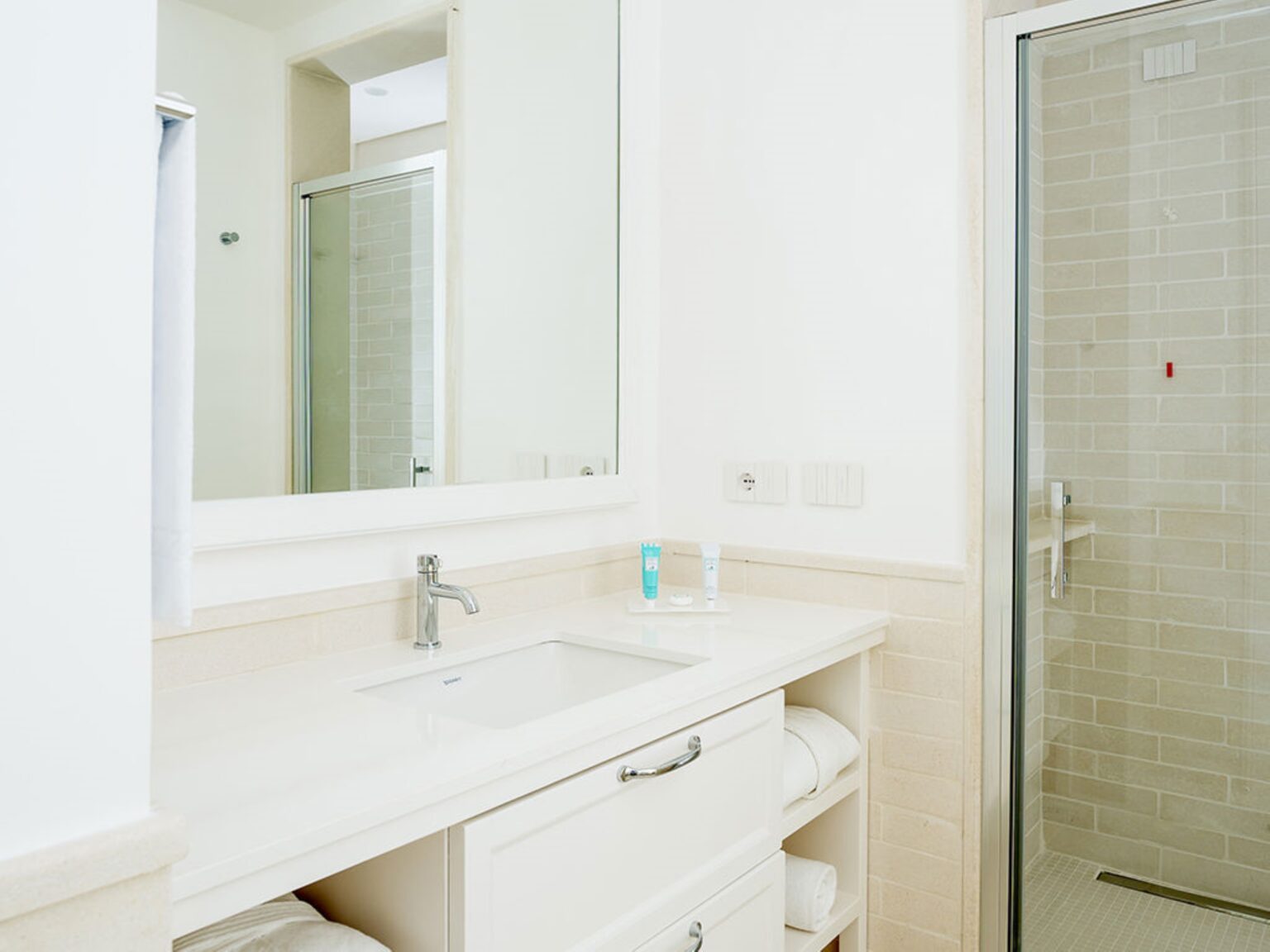 Modern bathroom interior featuring a sleek white sink, vanity with storage, and a glass shower enclosure. Natural light enhances the clean design, complemented by neutral tile and soft towels. Ideal for a relaxing and stylish space. | Only on SopranoVillas
