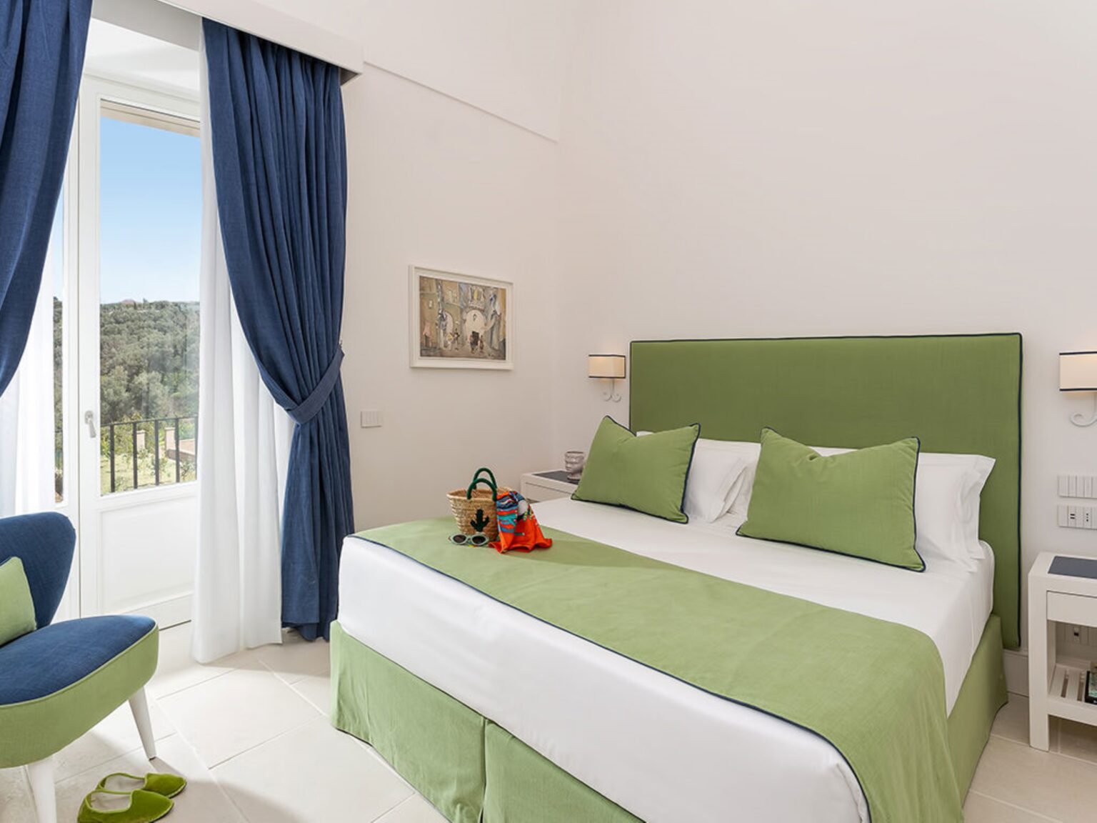 Cozy hotel room featuring a green accent wall, a comfortable double bed with matching green linens, and large windows with blue curtains. The room includes a small seating area, artwork on the wall, and a picturesque view of the outdoors. Ideal for a relaxing getaway. | Only on SopranoVillas