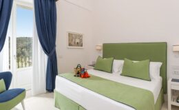 Cozy hotel room featuring a green accent wall, a comfortable double bed with matching green linens, and large windows with blue curtains. The room includes a small seating area, artwork on the wall, and a picturesque view of the outdoors. Ideal for a relaxing getaway.