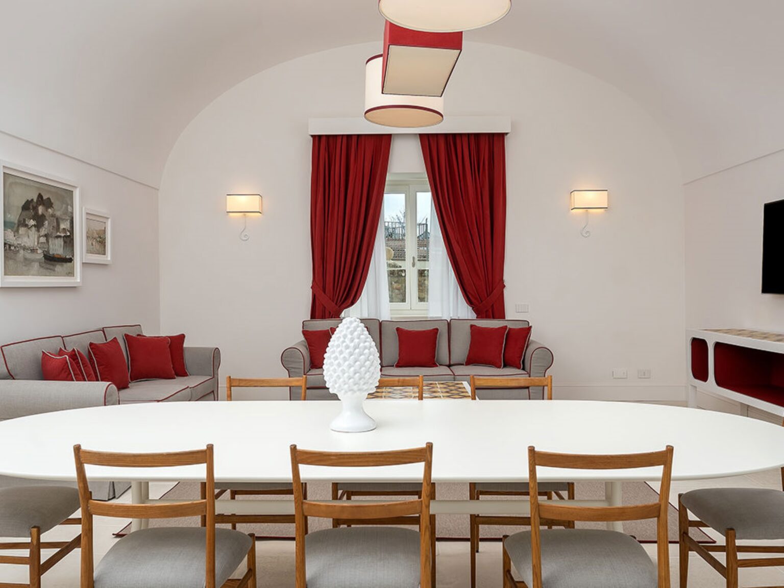 Modern living room and dining area featuring a white oval table surrounded by wooden chairs, a gray sofa with red cushions, and elegant red drapes. The space is illuminated by stylish light fixtures, creating a warm and inviting atmosphere. | Only on SopranoVillas