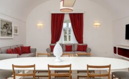 Modern living room and dining area featuring a white oval table surrounded by wooden chairs, a gray sofa with red cushions, and elegant red drapes. The space is illuminated by stylish light fixtures, creating a warm and inviting atmosphere.
