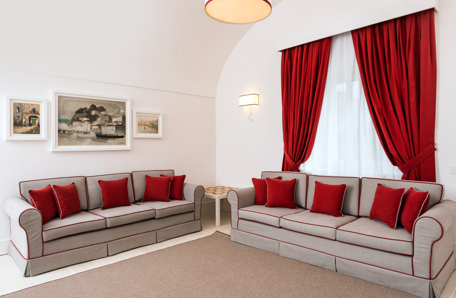 Modern living room featuring two gray sofas with red accent pillows, elegant red curtains, and framed artwork on the wall, creating a stylish and inviting atmosphere. | Only on SopranoVillas