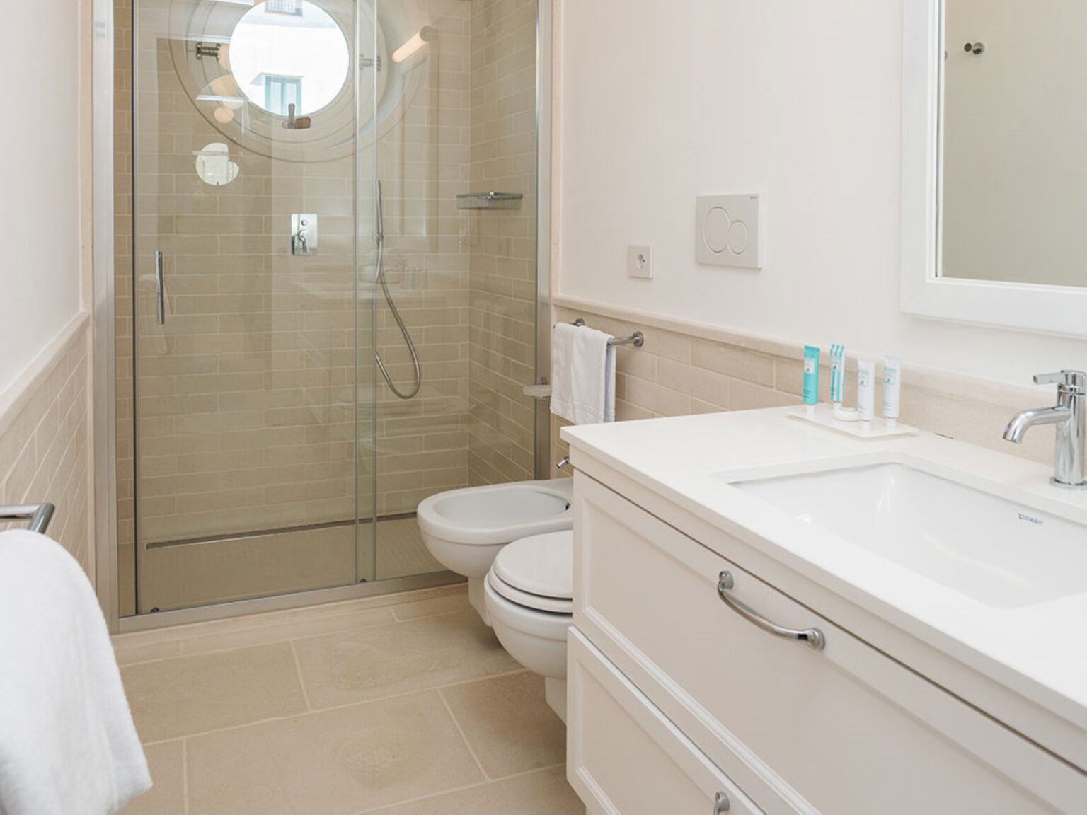 Modern bathroom featuring a glass shower enclosure, round window, white cabinetry, and a clean, minimalist design. The space includes a bidet and a sleek sink with toiletries arranged neatly on the countertop. Bright, neutral tones enhance the airy atmosphere. | Only on SopranoVillas