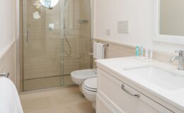 Modern bathroom featuring a glass shower enclosure, round window, white cabinetry, and a clean, minimalist design. The space includes a bidet and a sleek sink with toiletries arranged neatly on the countertop. Bright, neutral tones enhance the airy atmosphere.