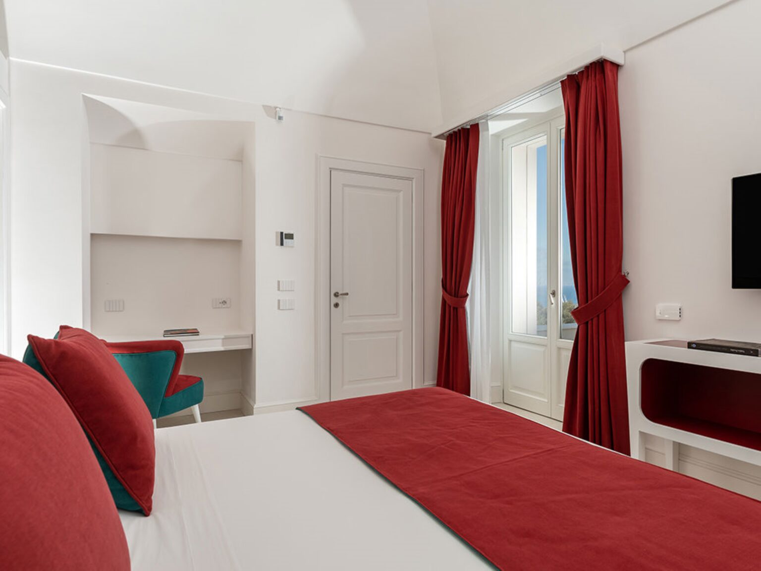Modern hotel room featuring a king-size bed with red accents, stylish curtains, a work desk, and a flat-screen TV, designed for comfort and relaxation. | Only on SopranoVillas