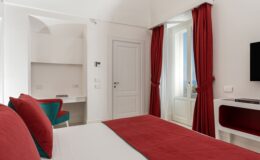 Modern hotel room featuring a king-size bed with red accents, stylish curtains, a work desk, and a flat-screen TV, designed for comfort and relaxation.