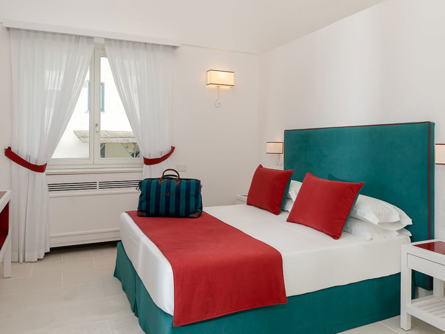 Modern hotel room featuring a king-size bed with red and teal accents, white linens, and decorative pillows. The room includes large windows with sheer white curtains, a stylish bedside table, and a colorful duffel bag placed on the bed. Ideal for travelers seeking a chic and comfortable stay. | Only on SopranoVillas