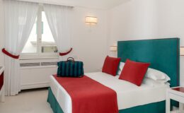 Modern hotel room featuring a king-size bed with red and teal accents, white linens, and decorative pillows. The room includes large windows with sheer white curtains, a stylish bedside table, and a colorful duffel bag placed on the bed. Ideal for travelers seeking a chic and comfortable stay.
