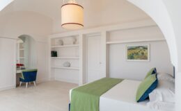 Modern bedroom interior featuring a white color scheme, a green-accented bed, a stylish pendant light, and a cozy workspace with a blue chair. The room includes built-in shelves and a door leading to another area, creating a spacious and inviting atmosphere. Ideal for relaxation and productivity.