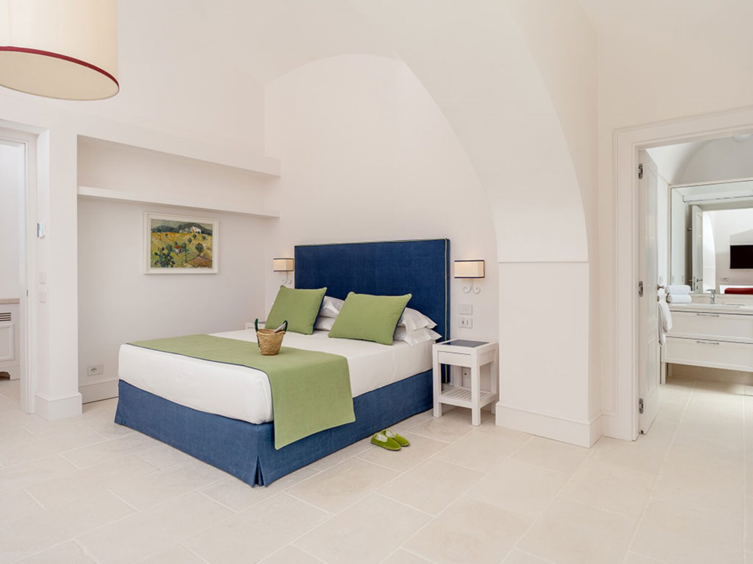 Modern hotel bedroom featuring a blue bed with green accents, minimalist decor, and natural light. The room includes a bedside table, artwork on the wall, and an open doorway leading to a stylish bathroom. Ideal for travelers seeking comfort and elegance. | Only on SopranoVillas