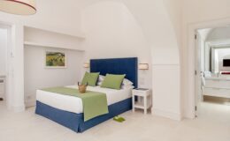Modern hotel bedroom featuring a blue bed with green accents, minimalist decor, and natural light. The room includes a bedside table, artwork on the wall, and an open doorway leading to a stylish bathroom. Ideal for travelers seeking comfort and elegance.