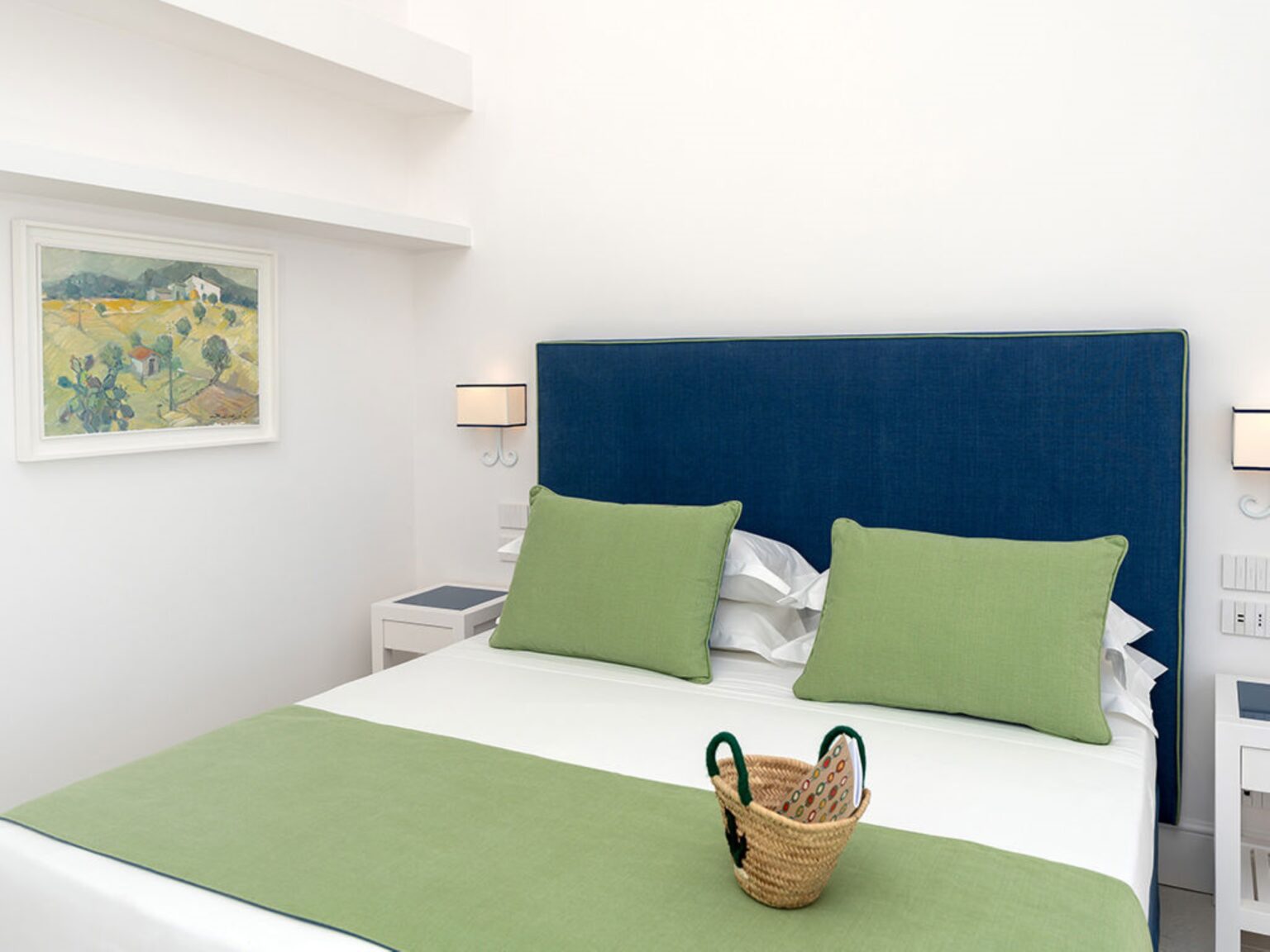 Modern bedroom featuring a blue upholstered headboard, green accent pillows, and a minimalist design. The room includes a cozy bed with white linens, a decorative basket, and a framed landscape artwork on the wall, creating a serene and inviting atmosphere. | Only on SopranoVillas