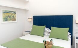 Modern bedroom featuring a blue upholstered headboard, green accent pillows, and a minimalist design. The room includes a cozy bed with white linens, a decorative basket, and a framed landscape artwork on the wall, creating a serene and inviting atmosphere.