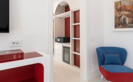 Interior view of a modern living space featuring an archway leading to a kitchenette with a microwave and shelves, complemented by a stylish blue chair and red accents. Ideal for showcasing contemporary home design and functionality.