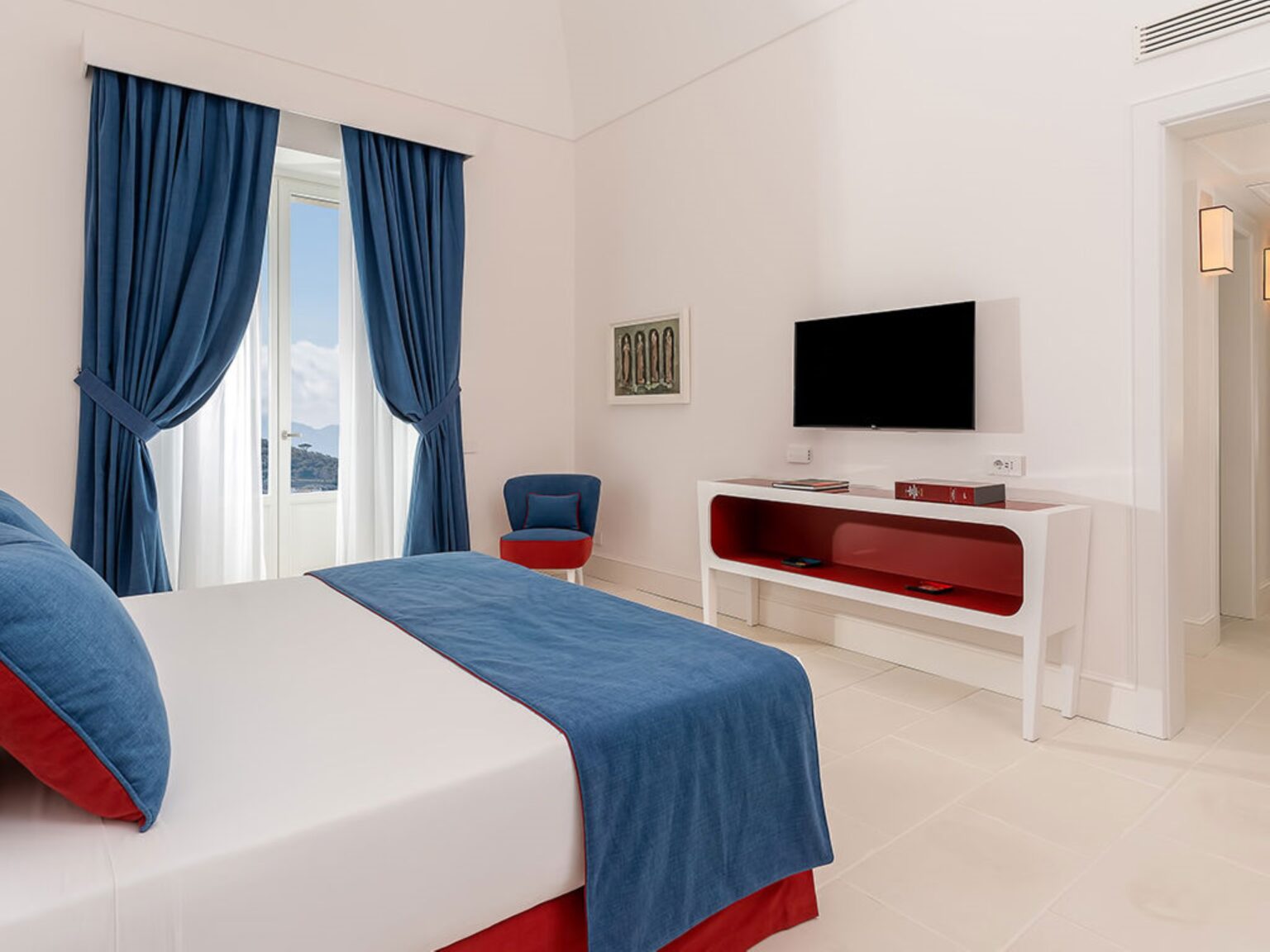 Modern hotel bedroom featuring a queen-sized bed with blue and red accents, a flat-screen TV mounted on a stylish white console, and large windows dressed with blue curtains, creating a bright and inviting atmosphere. | Only on SopranoVillas