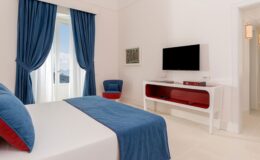 Modern hotel bedroom featuring a queen-sized bed with blue and red accents, a flat-screen TV mounted on a stylish white console, and large windows dressed with blue curtains, creating a bright and inviting atmosphere.
