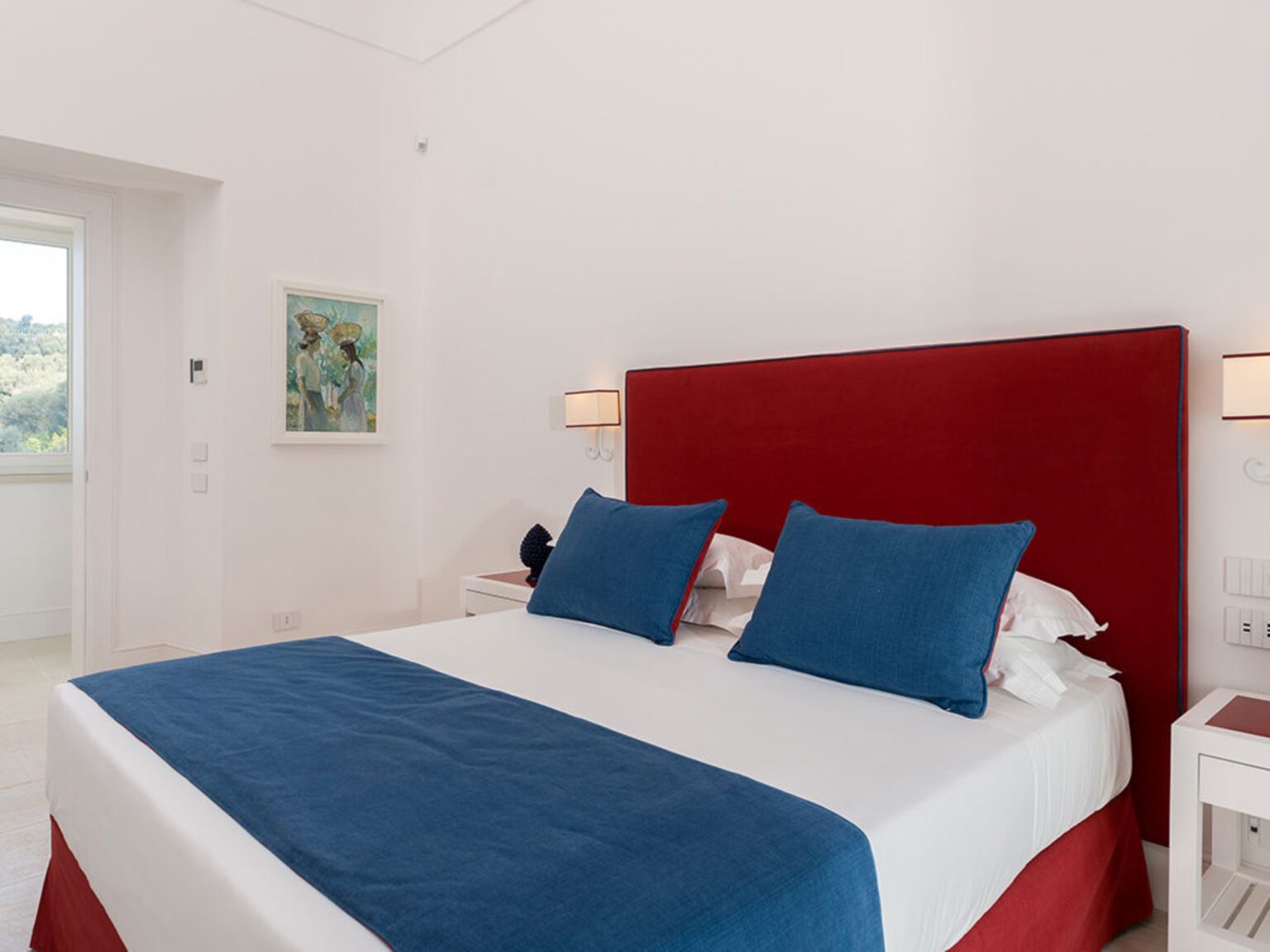 Modern bedroom featuring a red upholstered headboard, blue accent pillows, and a white bedspread. Natural light enters through a nearby window, enhancing the bright and airy atmosphere. Ideal for vacation rentals or hotel accommodations. | Only on SopranoVillas