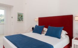 Modern bedroom featuring a red upholstered headboard, blue accent pillows, and a white bedspread. Natural light enters through a nearby window, enhancing the bright and airy atmosphere. Ideal for vacation rentals or hotel accommodations.