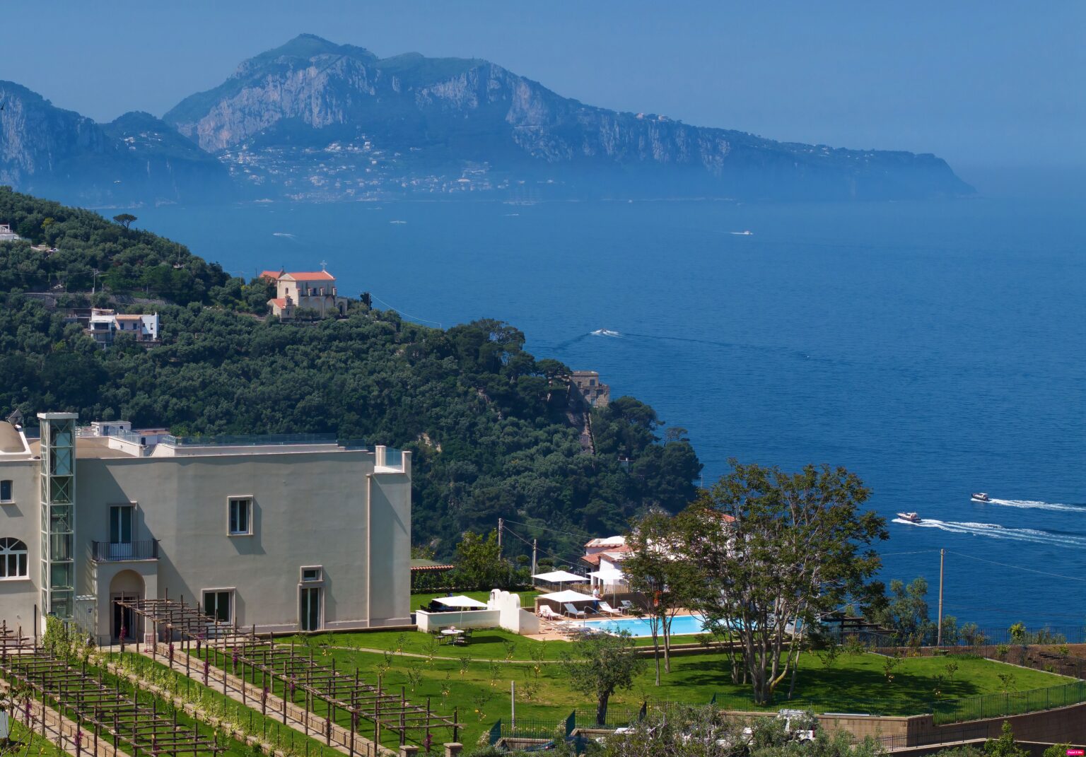 Coastal view of Capri, Italy, featuring lush greenery, a modern villa, and the serene blue Mediterranean Sea with distant mountains in the background. | Only on SopranoVillas