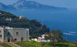 Coastal view of Capri, Italy, featuring lush greenery, a modern villa, and the serene blue Mediterranean Sea with distant mountains in the background.
