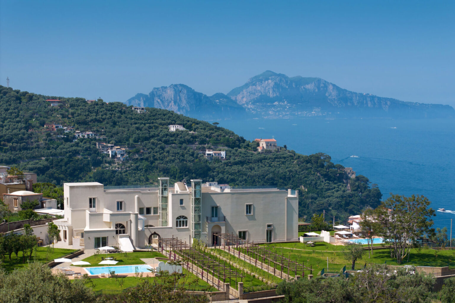 Luxury villa overlooking the sea with scenic mountains in the background, featuring lush gardens and swimming pools, ideal for a high-end getaway. | Only on SopranoVillas