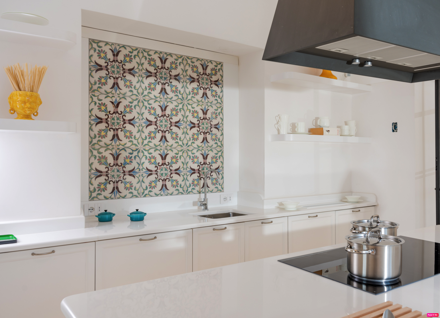 Modern kitchen interior featuring a stylish backsplash with floral pattern, stainless steel pots on a sleek countertop, and minimalist white cabinetry. Decorative items include a yellow vase with pasta and turquoise cookware, creating a vibrant and inviting atmosphere. | Only on SopranoVillas